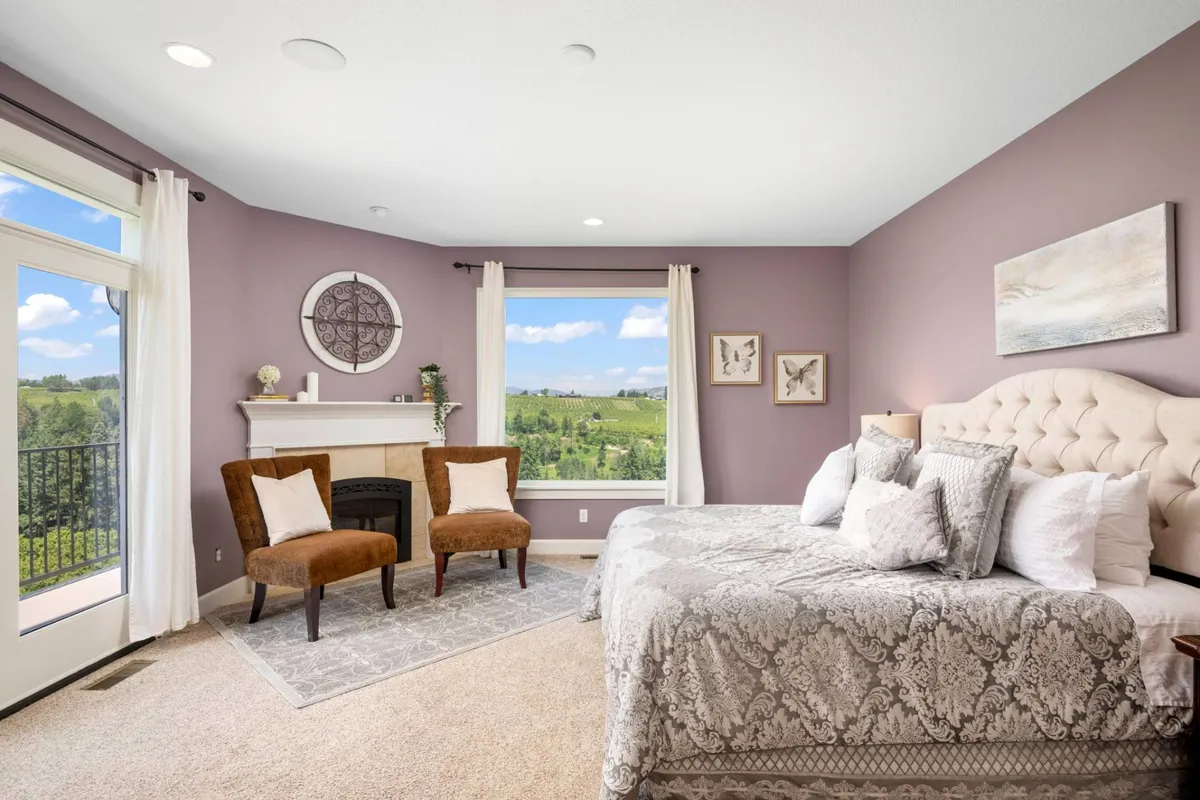 A serene bedroom featuring a plush bed with decorative pillows, comfortable chairs, a fireplace, and large windows that reveal idyllic views of the surrounding countryside. The soft linens and tranquil color palette create an inviting, restful ambiance.