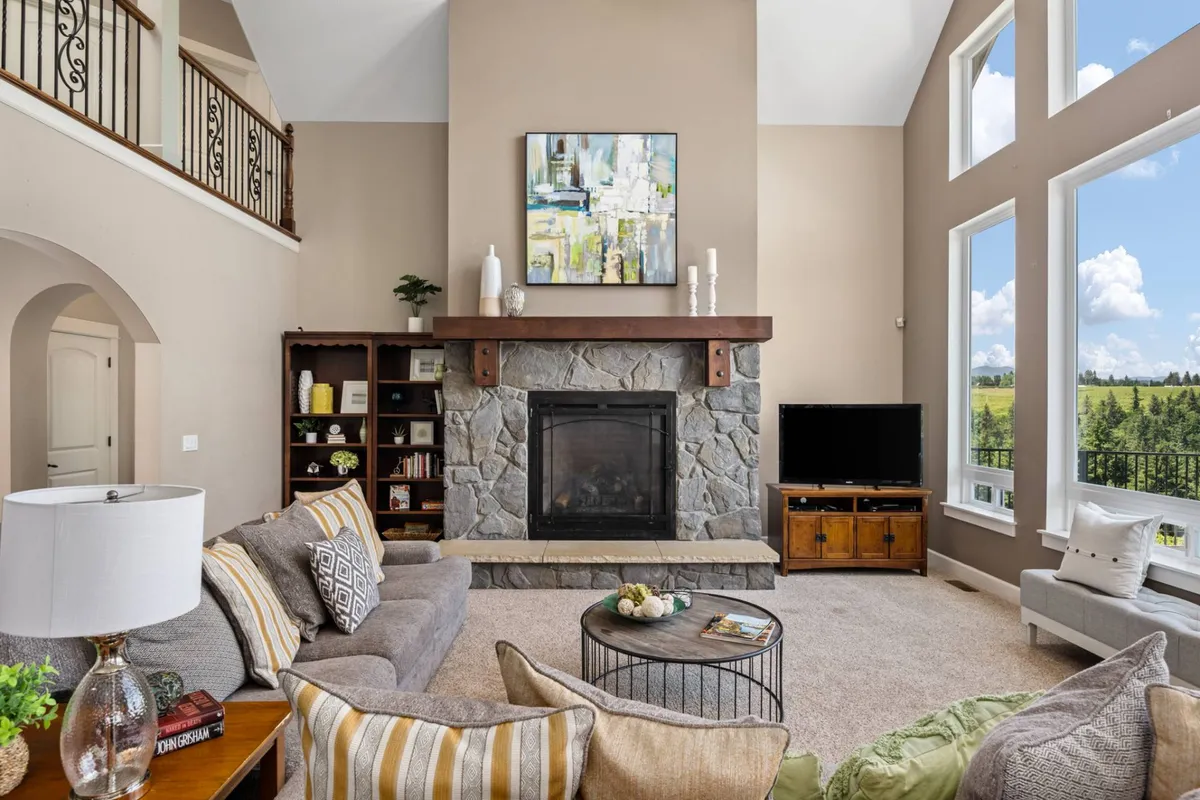 A welcoming living room with a striking stone fireplace as the focal point, framed by expansive windows that reveal the lush outdoors, and comfortable seating arranged for socializing or relaxing. The thoughtfully curated artwork above the fireplace and high-end furnishings hint at luxury and an eye for detail.