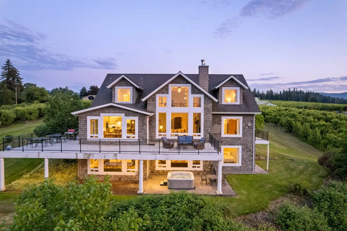 The hero image showcases the elegant exterior of RiverSong Country Chateau, featuring expansive windows that frame mountain views, a spacious outdoor deck perfect for gatherings, a bubbling private hot tub, and a beautifully landscaped yard. Visible amenities suggest space for outdoor dining, relaxation in the hot tub, and seamless indoor-outdoor living, all set against the tranquil backdrop of the Hood River Valley.