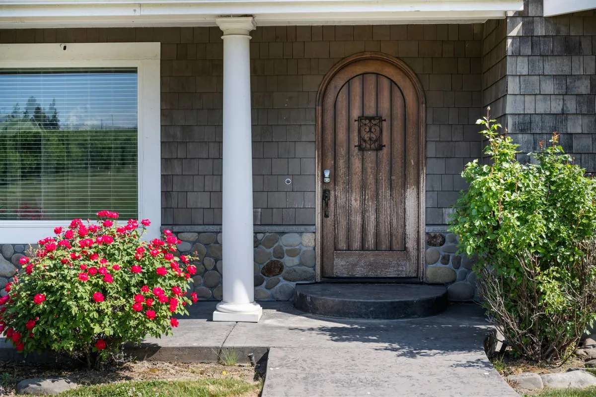 A charming exterior shot of the home's entrance with a sturdy wooden door, lush red rose bushes, a stone pathway, and elegant columns. The inviting garden and accessible entry signal the beginning of outdoor adventures, setting the tone for welcoming both guests and their dogs.