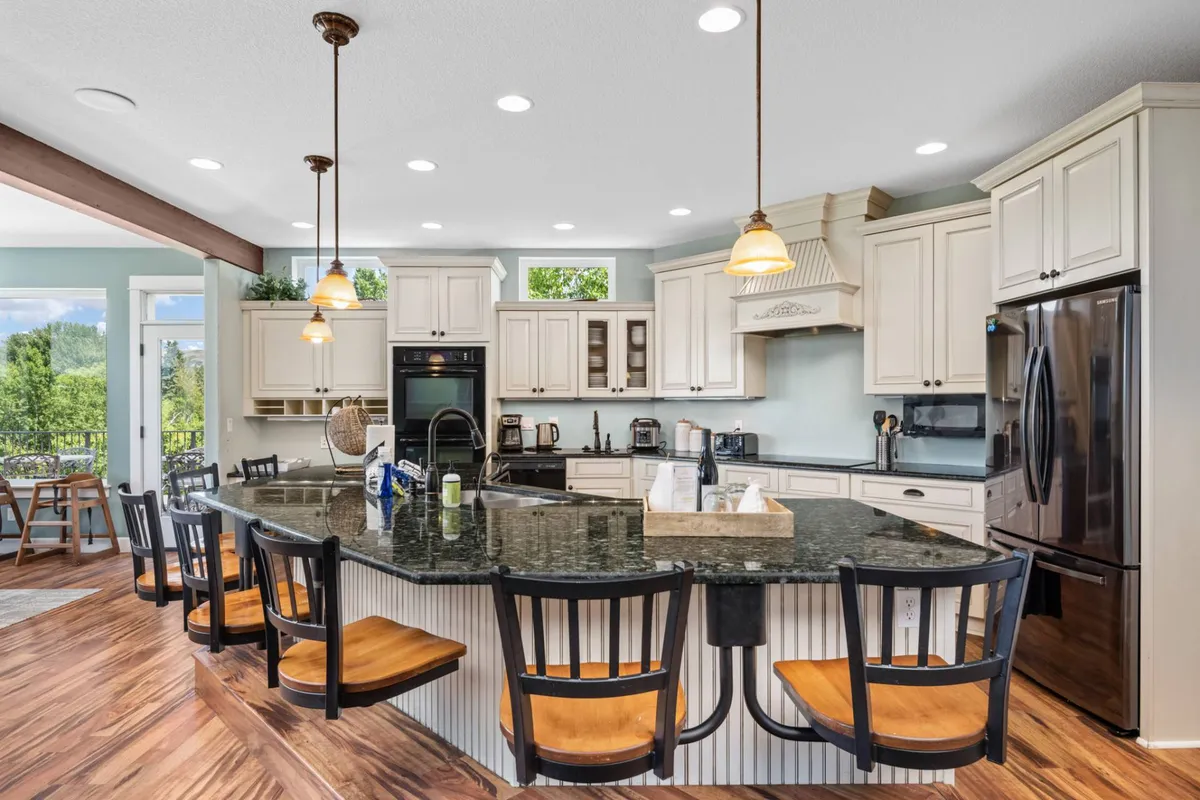 A spacious, well-lit gourmet kitchen featuring a large kitchen island with granite countertops, stainless steel high-end appliances, and pendant lighting overhead. The scene is styled for hosting, with space for group meal prep and casual socializing, evoking the heart of a home where recipes are shared and milestones celebrated.