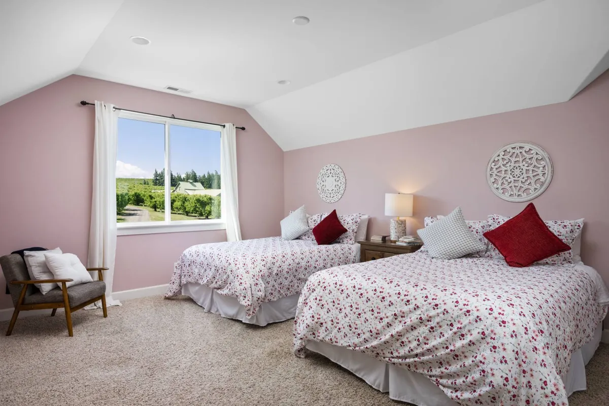 A comfortable bedroom with two beds adorned in floral bedding, a large window offering views of the orchard or countryside, decorative wall art, and a warm nightstand lamp. The layout is spacious and welcoming for families or groups.